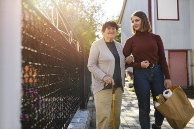 Na zdjeciu widzimy seniorkę z wolontariuszką niosącą reklamówkę z zakupami